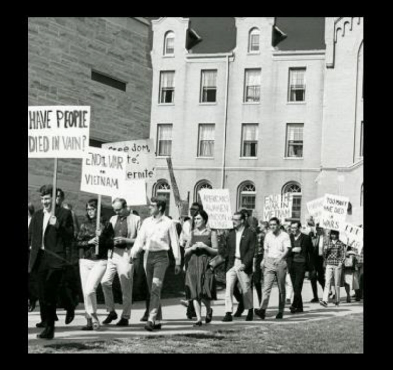 Photo of Students at NIU protesting the Vietnam War in 1965 by walking in a line and carrying various signs. 