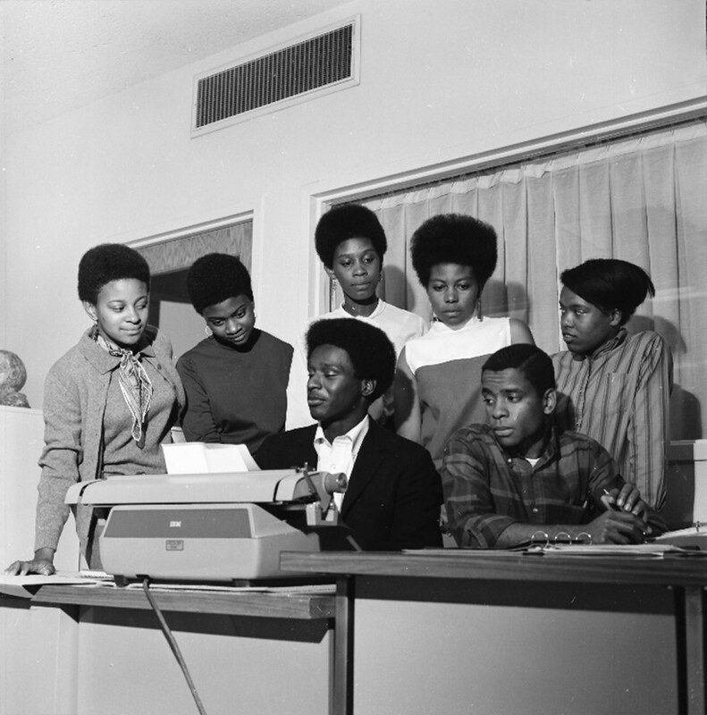 Photograph of 7 students in the African Students Association in the 1980's huddled around a typewriter. 
